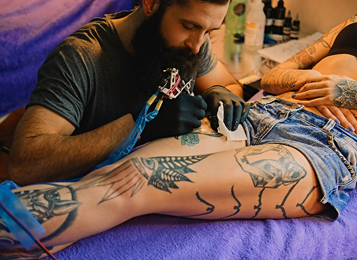 Tattoo artist in black gloves working on a client's leg tattoo, illustrating tattoo artists and moral grounds they decline jobs on.