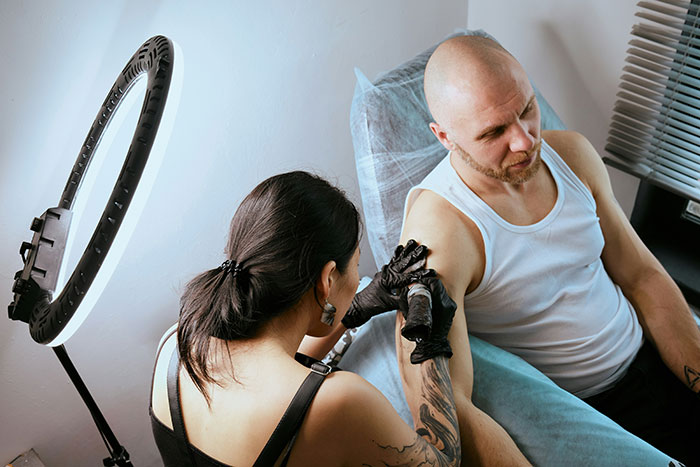 Tattoo artist wearing gloves using a machine to create a tattoo on a seated man’s upper arm under bright ring light.