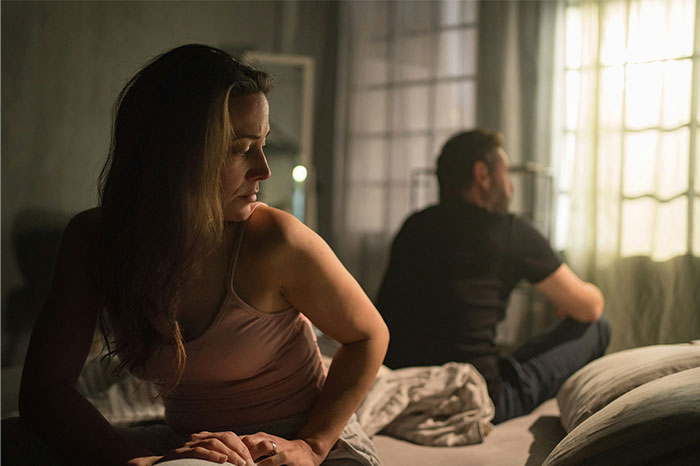 A distressed woman and man sitting back to back on a bed in a dimly lit room, showing tension and shock.