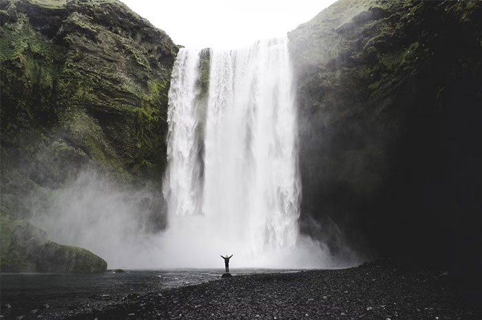 Person standing with arms raised near a large waterfall surrounded by rocky cliffs, illustrating deadly natural forces.