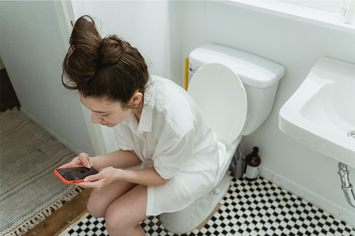 Woman sitting on toilet looking at phone in bathroom with checkered floor showing seemingly innocent things that are deadly risks.