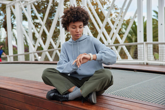 Young woman sitting cross-legged on a bench outdoors, checking her watch, symbolizing patience slowly disappearing.