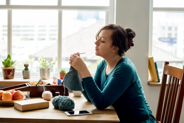Woman knitting by a sunlit window, surrounded by plants and yarn, reflecting patience from the good old days slowly disappearing.
