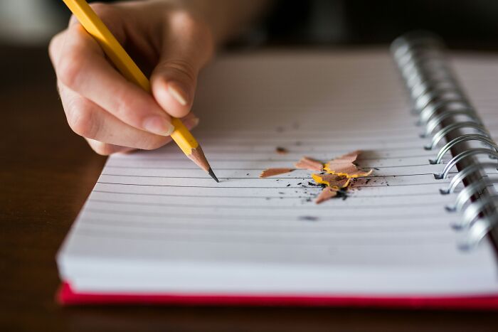Hand holding a yellow pencil writing on a notebook with pencil shavings on the page, symbolizing doctors and impostor patients.