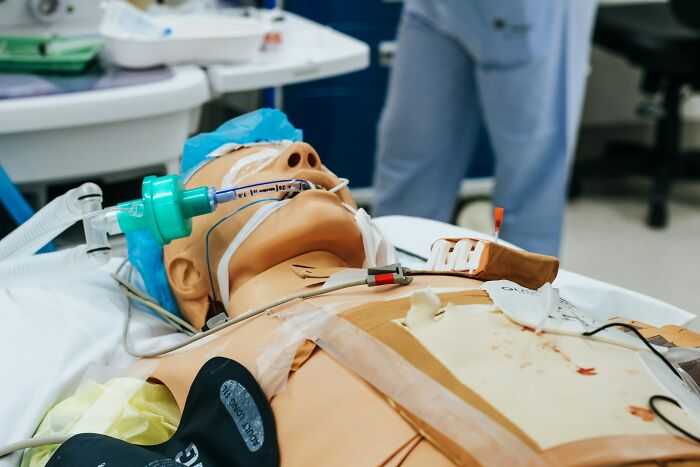 Medical training mannequin on hospital bed connected to breathing tube and monitoring devices for patient care simulation.