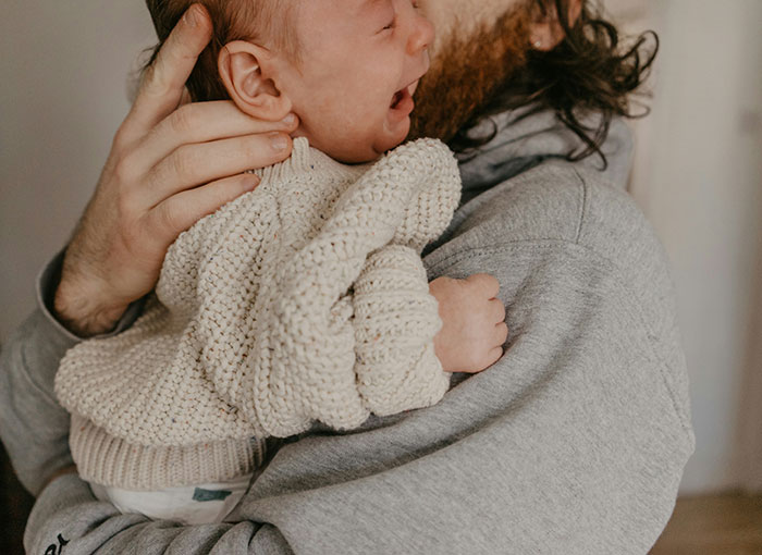 Man wearing a gray sweatshirt holding a crying baby wrapped in a knitted cream sweater indoors. - 11