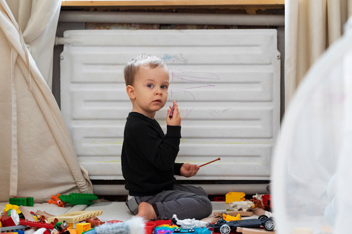 Toddler Karen banned from playing, sitting on floor with toys and drawing on a white radiator in a cluttered room. - 1