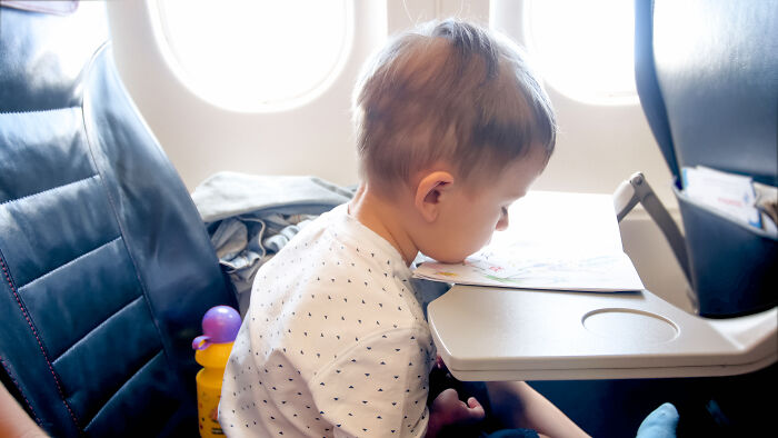 Toddler sitting on airplane seat using tray table for drawing during a public flight with a mom nearby managing the child.