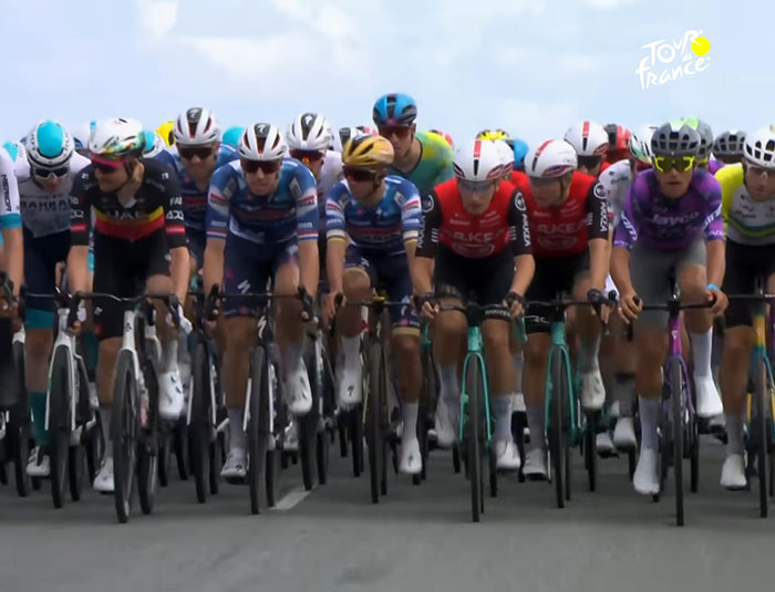 Tour de France cyclists ride closely in a group during a race, with fans controversially standing on graves nearby.