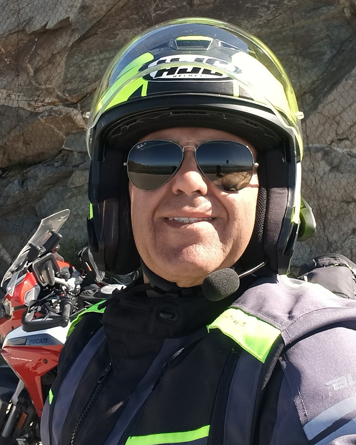 Motorcyclist wearing helmet and sunglasses taking a selfie near a rocky cliff with a red Ducati motorcycle parked nearby.