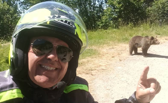 Tourist wearing helmet and sunglasses takes selfie near bear cub ignoring safety warnings in wooded area on sunny day.
