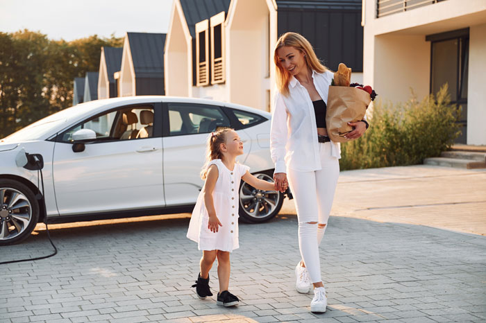 Woman and child walking past a white electric car parked in a residential driveway near a private parking space. Woman and child walking past a white electric car parked in a residential driveway near a private parking space.