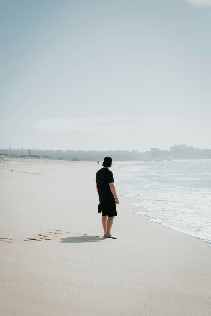 Person in black clothing standing alone on a sandy beach near the water, reflecting in a quiet moment for PIs weirdest cases. - 6