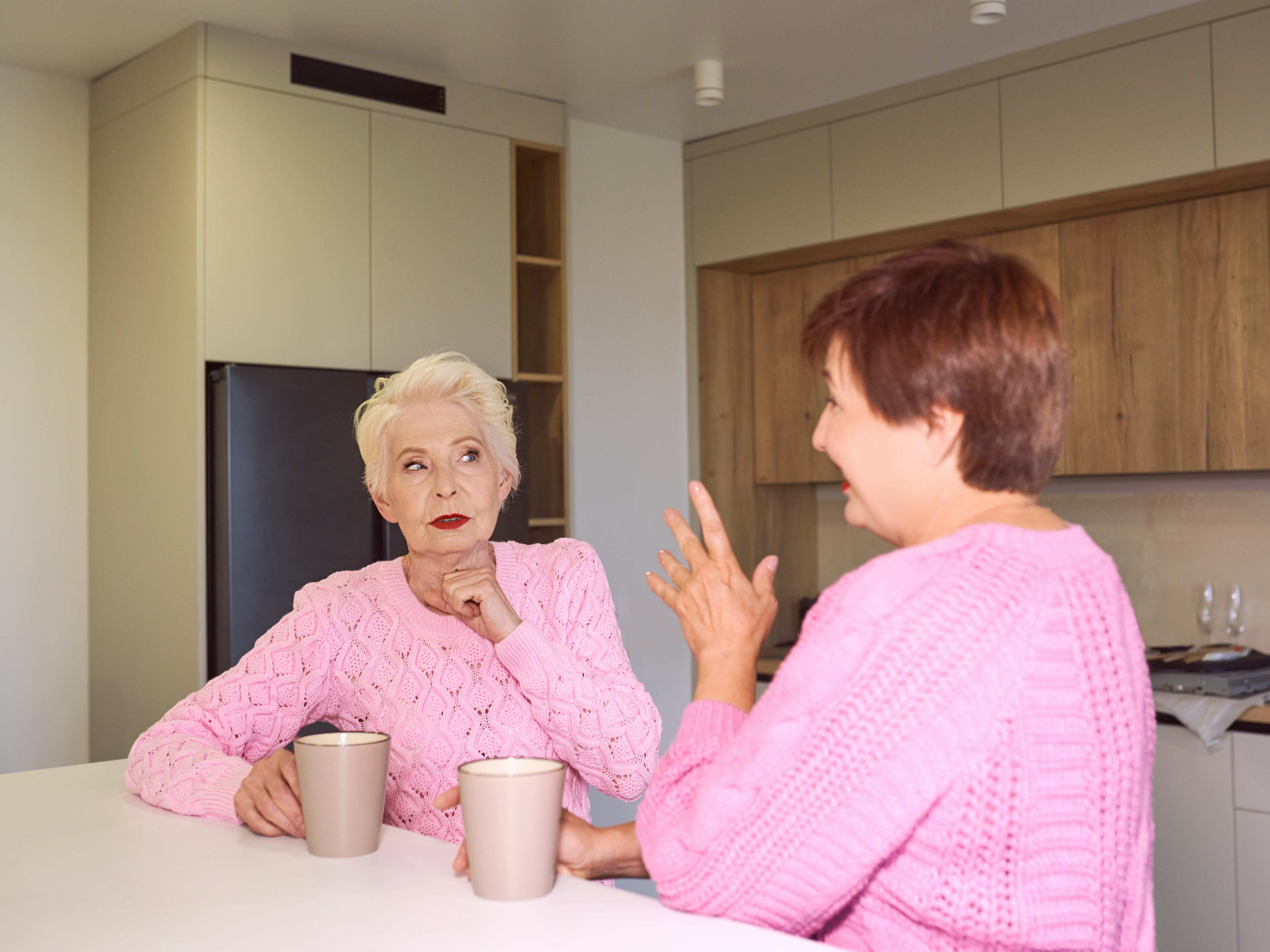 Two women in pink sweaters having a tense conversation in a kitchen, illustrating family drama over inherited house.