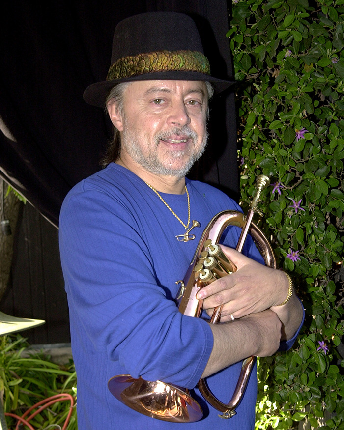 Jazz musician smiling and holding a brass instrument, wearing a black hat and blue shirt in an outdoor setting. - 1