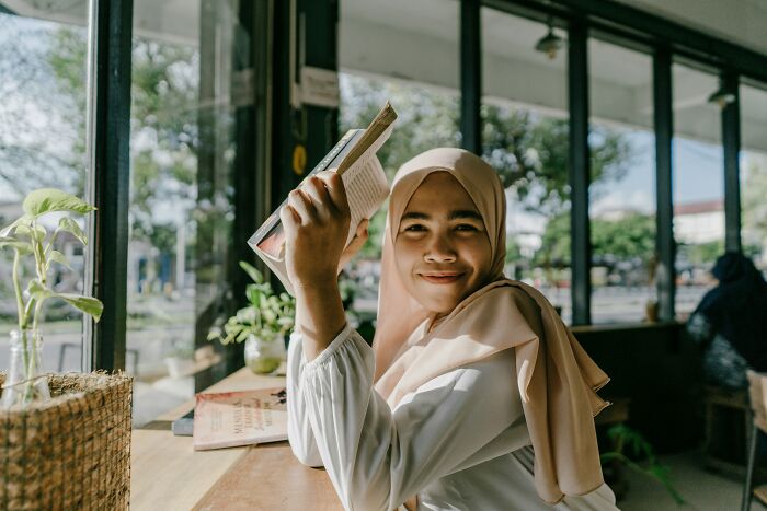 Young woman wearing a hijab holding a book and smiling, sitting by a window in a cafe discussing religious myths.