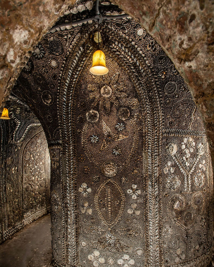 Intricate underground grotto walls covered in millions of seashells forming detailed patterns under warm lighting.