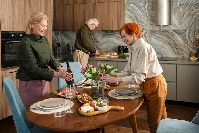 Two women setting a dining table in a modern kitchen with an unhinged family member wedding in the background. - 30