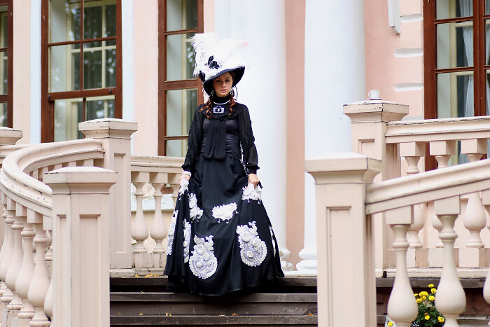 Person dressed in vintage black and white attire with large feathered hat, standing on mansion steps, unhinged family member wedding scene. - 23