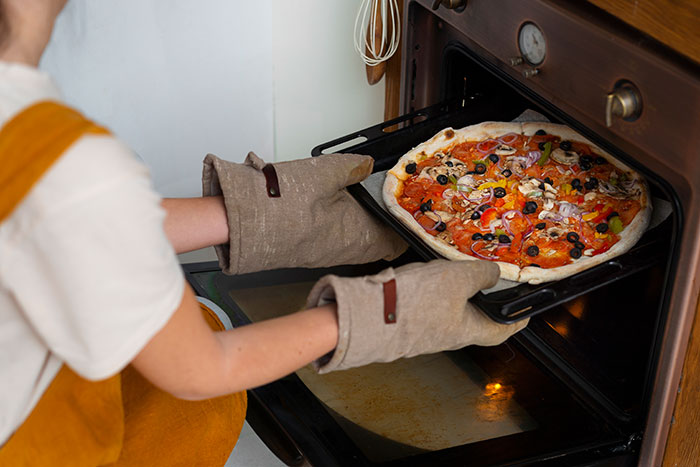Person using oven mitts to take a freshly baked pizza out of the oven, demonstrating life hacks that improve quality.