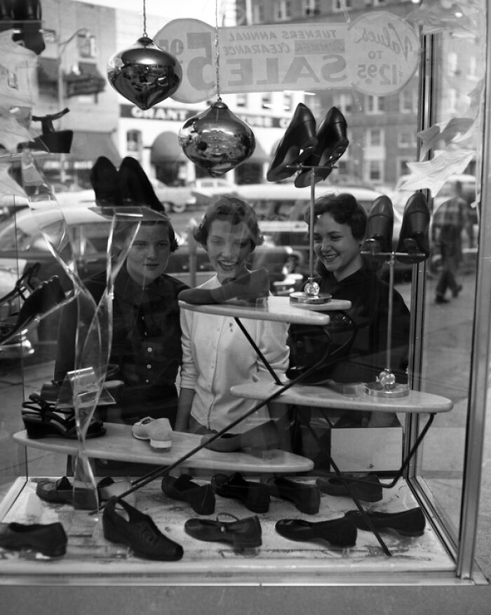 Three women looking at shoes displayed in a store window, capturing daily life across the globe in the 50s.