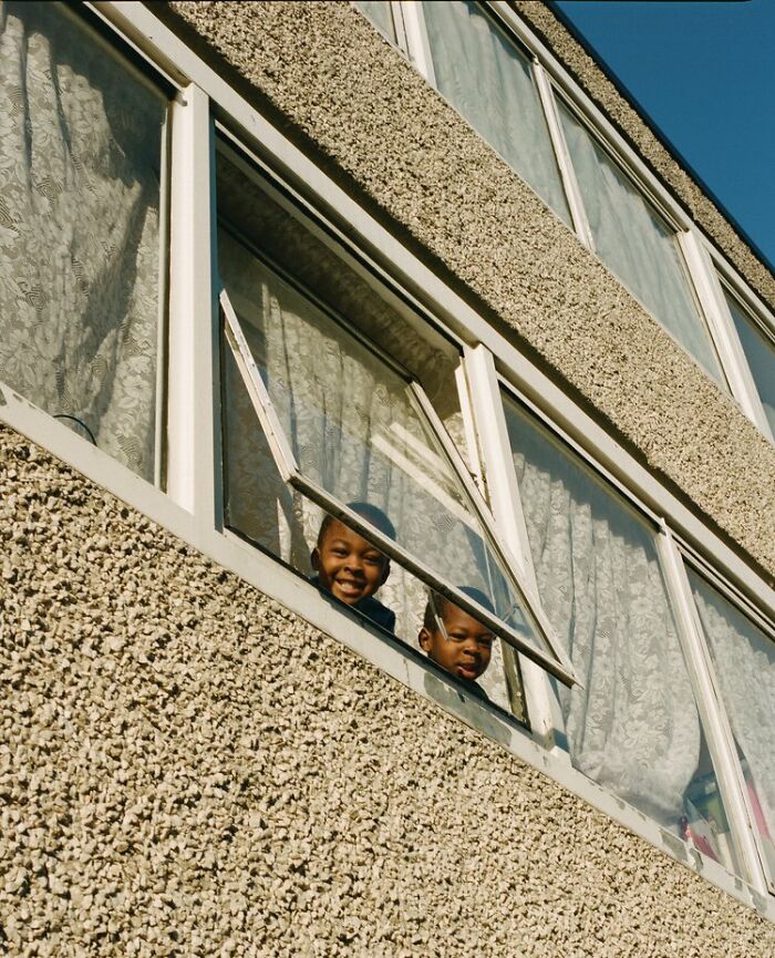 Two children smiling and looking out an open window of a textured building in London street photography by Nico Froehlich.