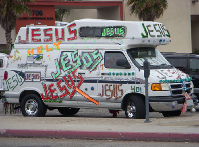 White van covered in oversized colorful text and decorations as an outrageous car mod parked on a city street.