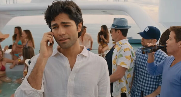 Man in white shirt talking on phone at a party with hard to love TV show characters in the background - 25