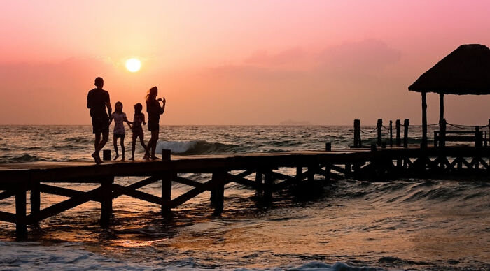 Family walking on a pier at sunset enjoying a vacation destination poll by the ocean with a pink sky background.