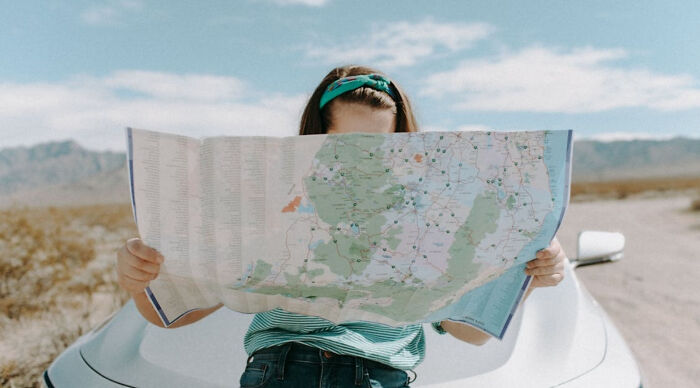 Person holding a map while sitting on a car in a desert setting, planning a vacation destination poll trip