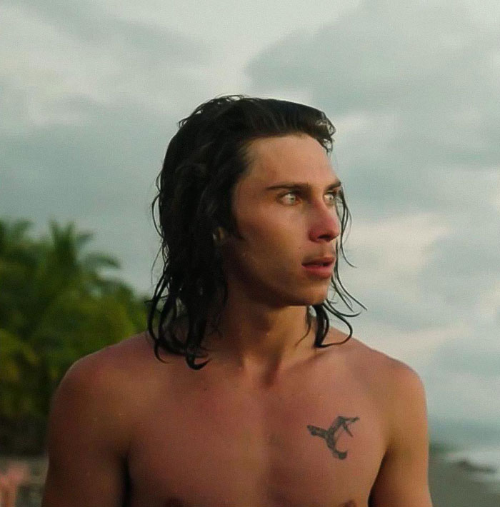 21YO daredevil with wet hair standing shirtless on beach, preparing for record-breaking dive during golden hour.