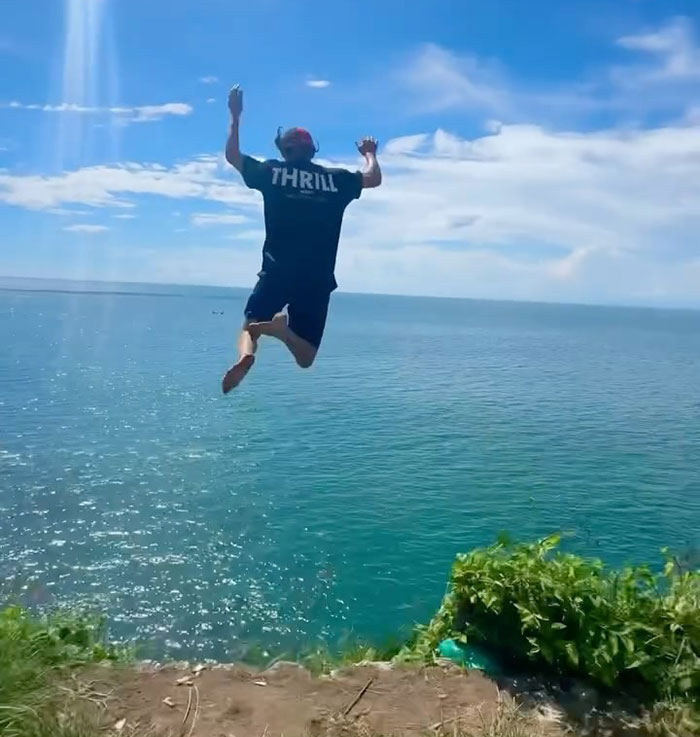 Young daredevil jumping off cliff into ocean attempting record-breaking dive on a sunny day with clear skies.