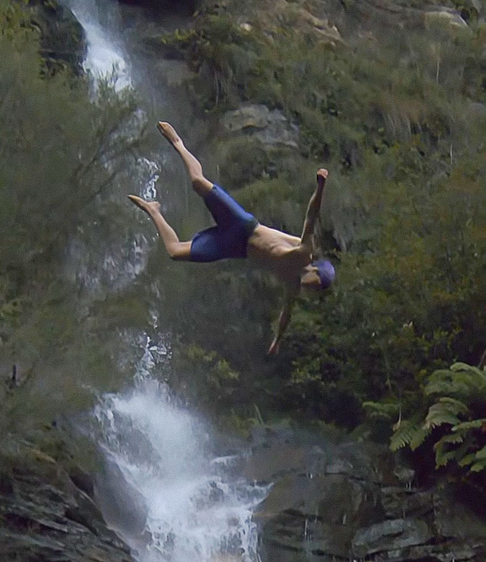Young daredevil mid-air during a record-breaking dive attempt near a rocky waterfall in a forested area.