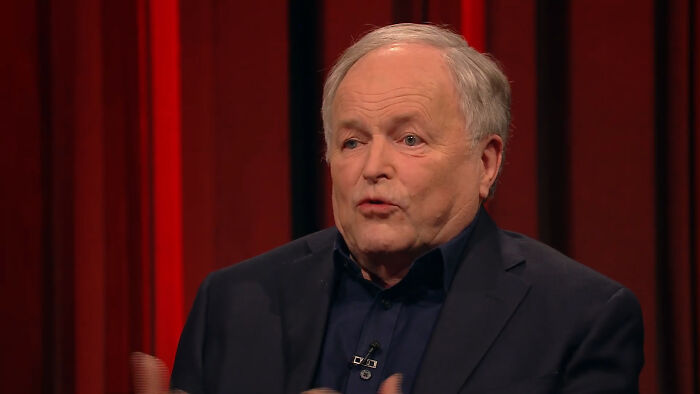 Older man in dark suit speaking on a red curtain stage, illustrating celebrities many people love but can't stand.