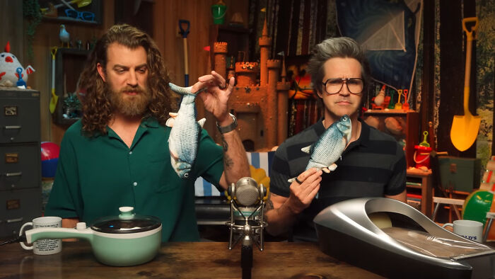 Two people share unsubscribed YouTubers, holding fish plushies while sitting at a table with various objects in a studio setting.