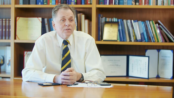 Middle-aged man in a white shirt and striped tie sitting at a desk in an office with books discussing historical events.