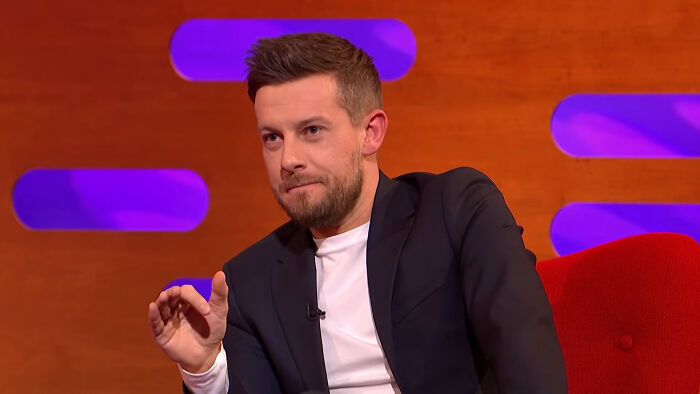Man in a dark suit sitting on a red chair during a TV show discussing celebrities that many love but others can't stand