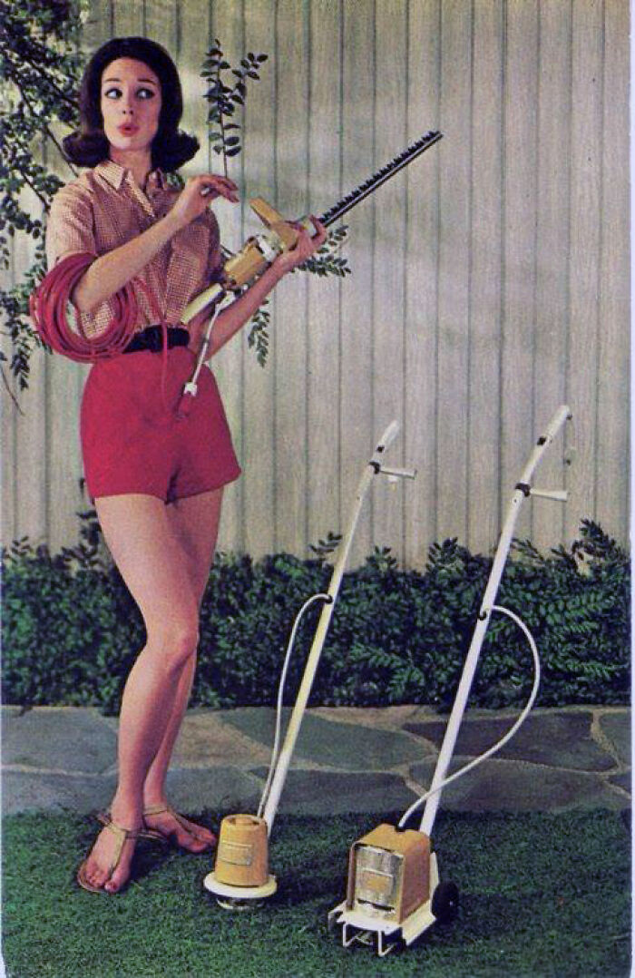 Woman in vintage outfit holding an old electric hedge trimmer near two vintage lawn mowers on grass lawn.