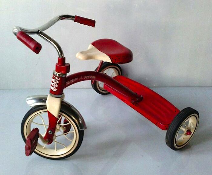 Vintage red tricycle with white and red seat, metal fenders, and three black and white wheels on a plain background.