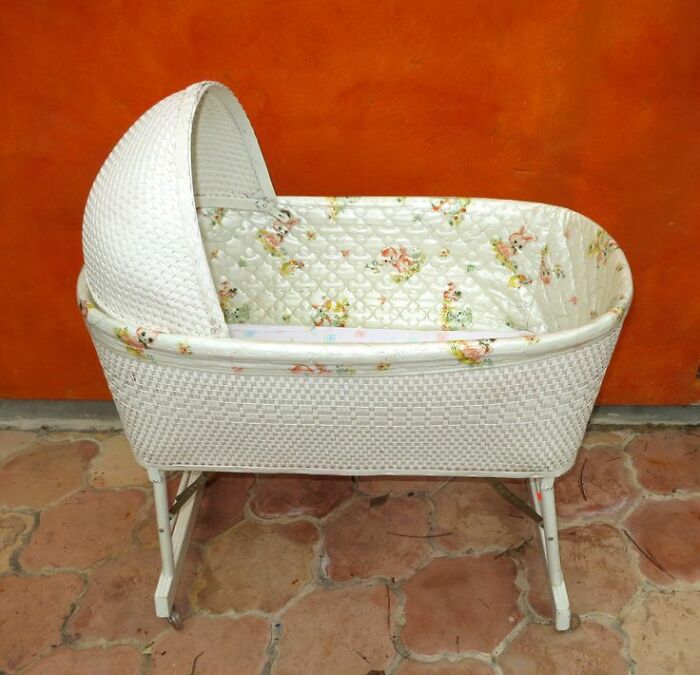 White vintage wicker bassinet with floral lining standing on tiled floor against orange wall.