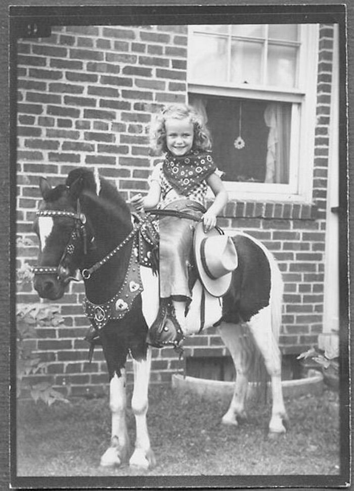 Vintage black and white photo of a child in cowboy attire sitting on a decorated pony in front of a brick house.