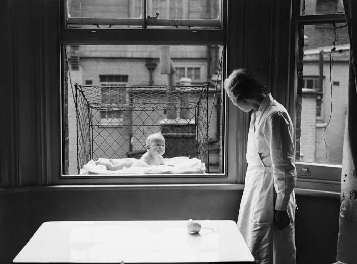 Vintage black and white photo of a baby in a weird invention outdoor window crib while a woman watches inside a room.