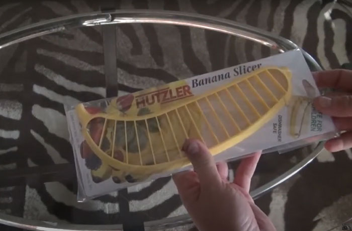 Hands holding a packaged weird invention, a yellow plastic banana slicer, over a glass table with a zebra print rug beneath.
