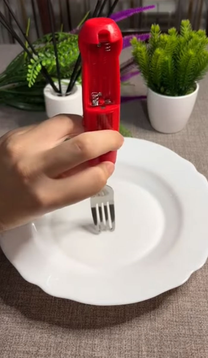 Hand holding a weird invention fork with red handle over a white plate on a table with green plants.