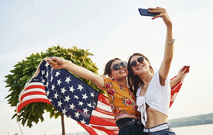 Two young women wearing sunglasses take a selfie outdoors holding an American flag, highlighting things non-Americans find weird. - 7