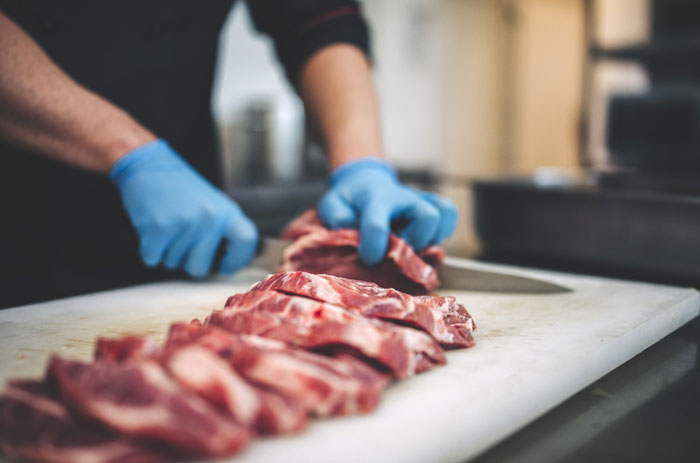 Person wearing blue gloves slicing raw meat, illustrating American foods banned in other countries due to regulations.