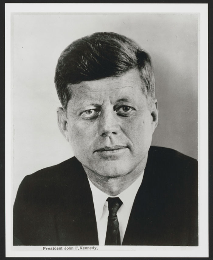Black and white close-up portrait of US president John F. Kennedy, linked to the highest IQ among presidents, wearing a suit and tie.