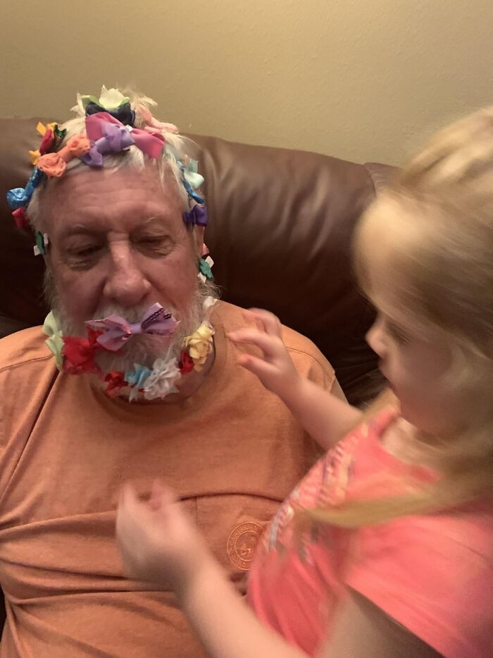 Elderly man adorned with colorful bows on his face and hair, engaging in a wholesome moment with a young child.