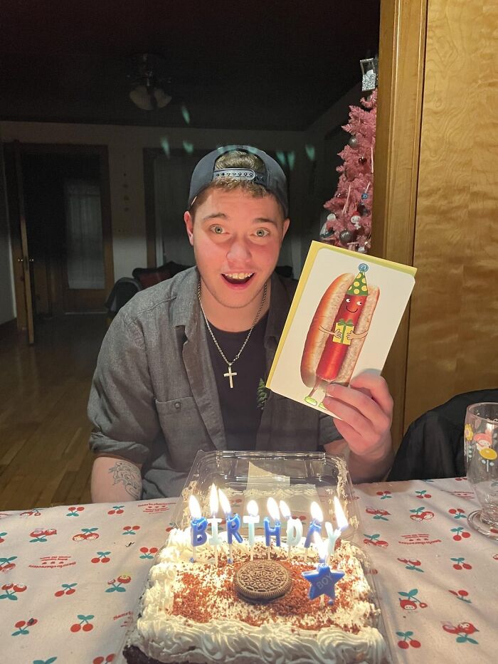 Young man smiling excitedly at birthday cake with lit candles, holding a greeting card, showcasing wholesome people and animals.