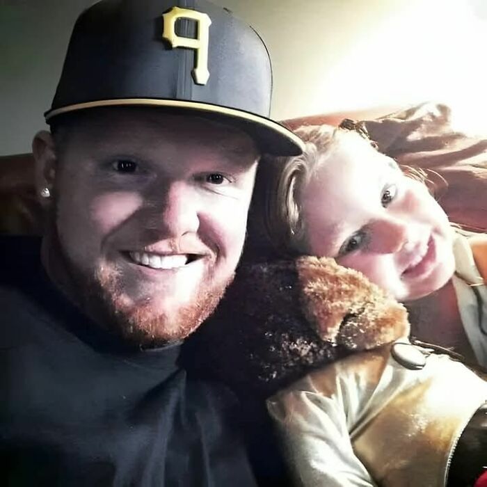Man wearing a black cap smiling with a young girl resting on a teddy bear, capturing a wholesome and amazing moment.